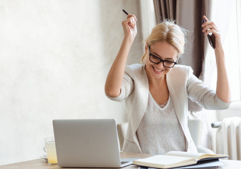 A photo of a female entrepreneur by a laptop with arms raised as she is pursuing her goals with a winning mindset