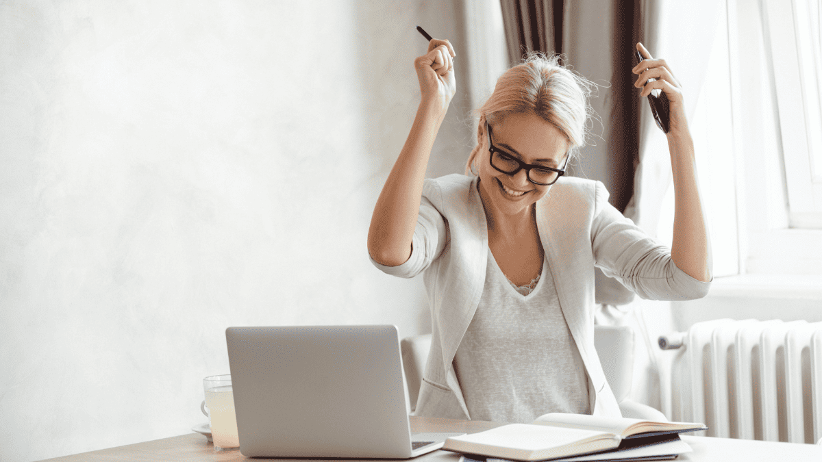 A photo of a female entrepreneur by a laptop with arms raised as she is pursuing her goals with a winning mindset